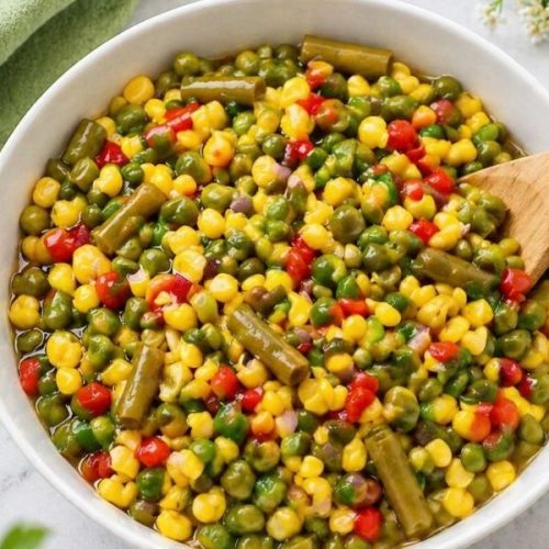 Fresh vegetable salad with mixed peas, corn, green beans, and peppers served in a white bowl on a light surface with a wooden spoon