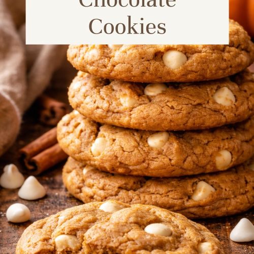 Stack of soft pumpkin white chocolate cookies filled with white chocolate chips and surrounded by cinnamon sticks and fall decor.