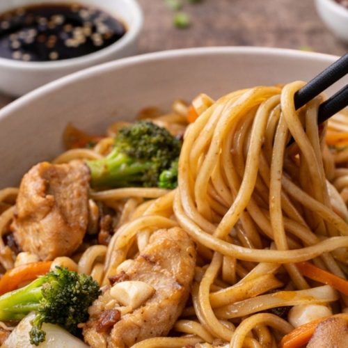 Close-up of chicken lo mein with chopsticks lifting noodles, mixed with sliced chicken, broccoli, carrots, mushrooms, and onions, with small bowls of sauce in the background.